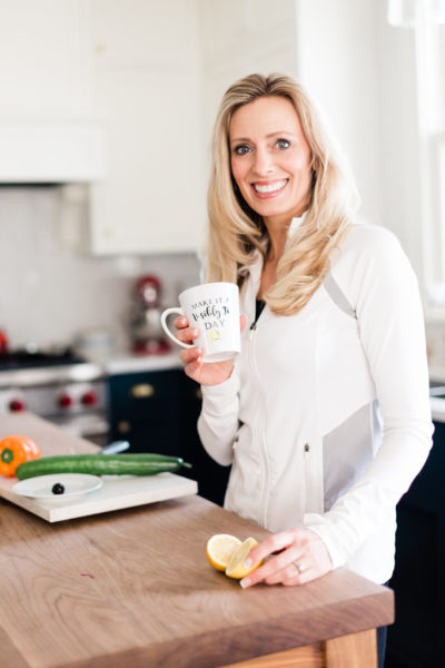 Wendie Pett smiling in the kitchen over fresh vegetables holding a "Make It A Visibly Fit™ Day" mug.