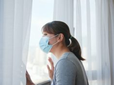 Woman with a surgical mask looking out a window with curtains to represent how to get Visibly Fit™ with Wendie Pett, ND even in the pandemic quarantine