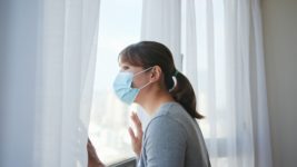 Woman with a surgical mask looking out a window with curtains to represent how to get Visibly Fit™ with Wendie Pett, ND even in the pandemic quarantine